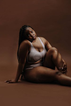 Porträt einer jungen Afrikanerin, die in Unterwäsche vor braunem Hintergrund in die Kamera blickt