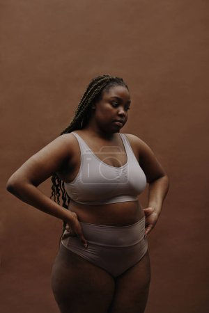 Porträt einer jungen Afrikanerin mit dicker Gestalt, die in Unterwäsche vor braunem Hintergrund steht