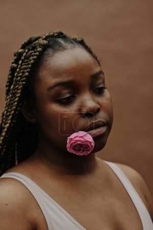 Photo for Portrait of African young woman with blank expression holding pink flower in her mouth - Royalty Free Image