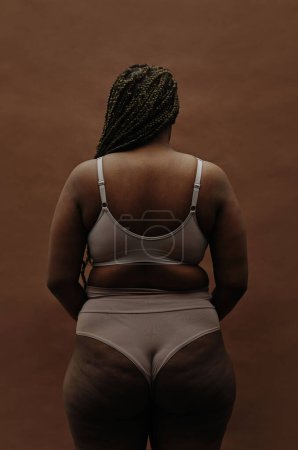 Rückansicht eines afrikanischen Mädchens mit dicker Figur, das in Unterwäsche vor braunem Hintergrund steht