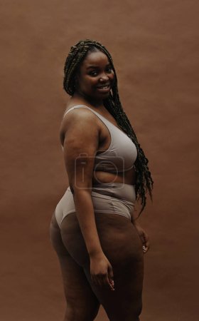 Porträt einer jungen Afrikanerin mit dicker Figur, die isoliert vor braunem Hintergrund in die Kamera lächelt