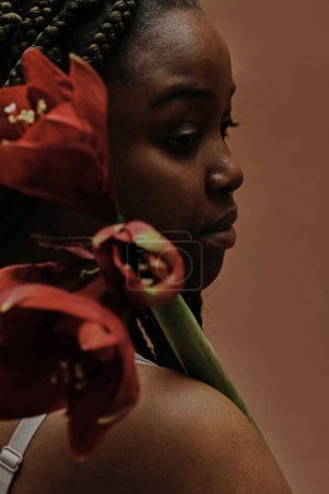 Photo for African girl looking through her shoulder holding big red flower isolated on brown background - Royalty Free Image