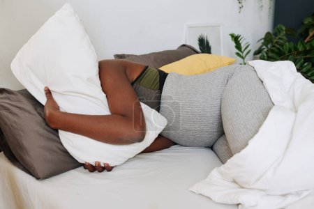 Foto de Mujer embarazada africana abrazando almohada y sufriendo de dolor acostado en la cama durante el parto - Imagen libre de derechos