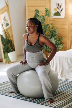 Chica embarazada sentada en la pelota de fitness y concentrándose con los ojos cerrados durante las contracciones