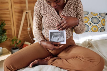 Photo for African pregnant girl looking at 3d ultrasound of her baby while sitting on bed in bedroom - Royalty Free Image