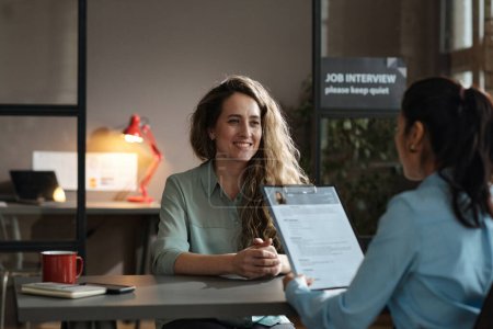 Foto de Mujer joven sentada en la mesa y sonriendo mientras el gerente examina su currículum durante la entrevista de trabajo - Imagen libre de derechos