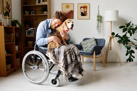 Foto de Joven barbudo abrazándose con su precioso perro mientras está sentado en silla de ruedas en la sala de estar - Imagen libre de derechos