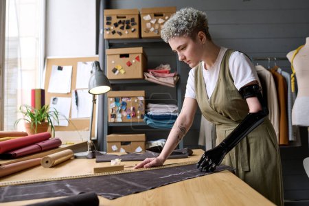 Young female tailor with black arm prosthesis standing by table in workshop or studio of modern fashion and creating new clothes