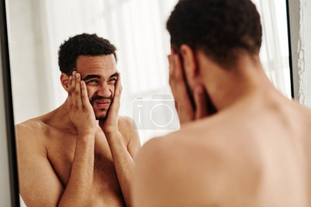 Photo for Young man holding his head in hands and looking at mirror with displeasure - Royalty Free Image
