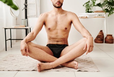 Téléchargez les photos : Gros plan d'un jeune homme torse nu assis sur un tapis en position lotus et faisant du yoga dans la salle de bain - en image libre de droit
