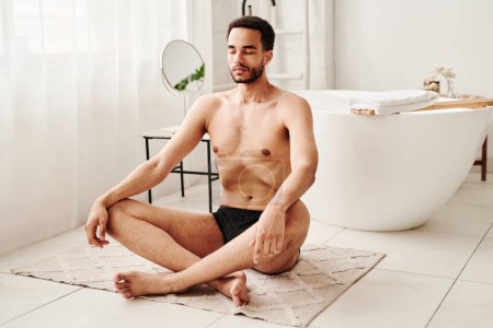 Téléchargez les photos : Jeune homme torse nu assis sur le tapis en position lotus et respirant profondément pendant la méditation dans la salle de bain - en image libre de droit