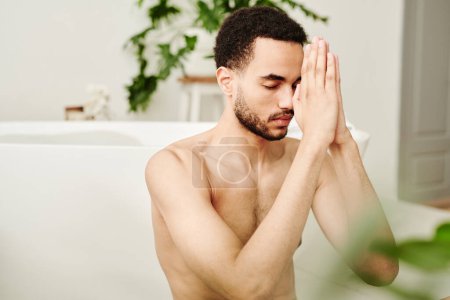 Téléchargez les photos : Jeune homme torse nu se concentrant pendant la méditation tout en étant assis dans la salle de bain - en image libre de droit