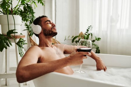 Photo for Young mixed race man listening to music in wireless headphones and drinking wine while relaxing in bath - Royalty Free Image
