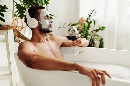 Photo for Young man enjoying relaxed time in bathroom, he listening to music in wireless headphones and enjoying the glass of red wine - Royalty Free Image