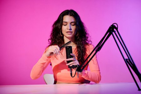 Téléchargez les photos : Jeune influenceuse blanche élégante assise au bureau dans un studio enregistrant des sons faits avec un peigne pour la vidéo ASMR, lumière rose néon - en image libre de droit