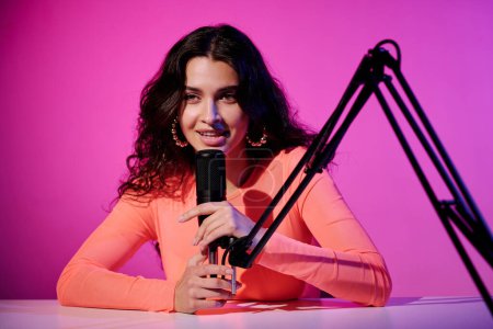 Hermosa joven influencer sentada en el escritorio en el estudio en luz de neón rosa susurrando en el micrófono para el video ASMR