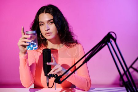 Joven blogger sentada en estudio en grabación de luz de neón rosa suena sacudiendo frasco con pastillas para ASMR vlog