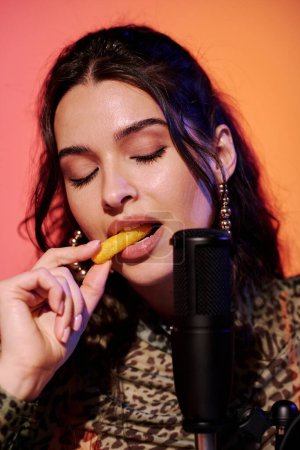 Téléchargez les photos : Attrayant jeune femme avec les yeux fermés assis en studio en rose et jaune néon lumière enregistrement maïs bouffées snacks manger des sons pour ASMR vlog - en image libre de droit