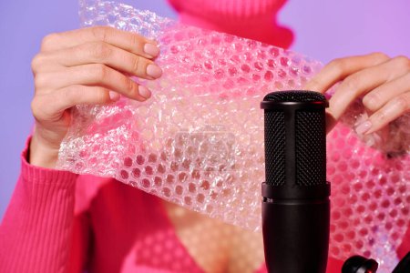 Unrecognizable young female influencer wearing pink clothes popping bubble wrap on microphone for ASMR content