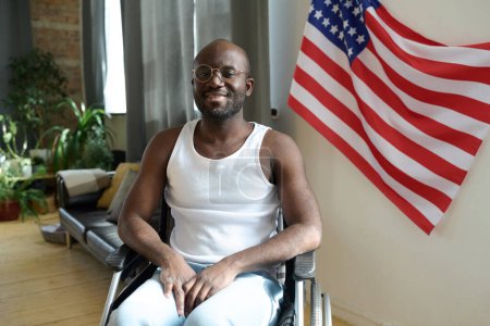 Portrait of African American man smiling at camera while sitting in wheelchair against the American flag on wall in the room