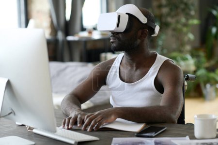 Photo for African young man in virtual reality goggles sitting at table in front of computer monitor and typing on keyboard - Royalty Free Image