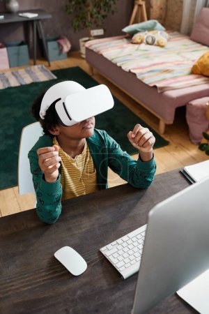 Photo for High angle view of African little boy sitting at table in front of computer and using VR goggles to play virtual reality game - Royalty Free Image