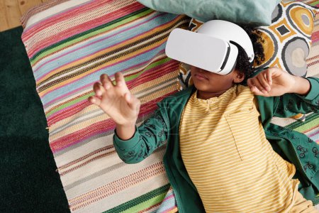 Photo for High angle view of African boy relaxing on sofa in VR goggles watching virtual reality movie and gesturing - Royalty Free Image