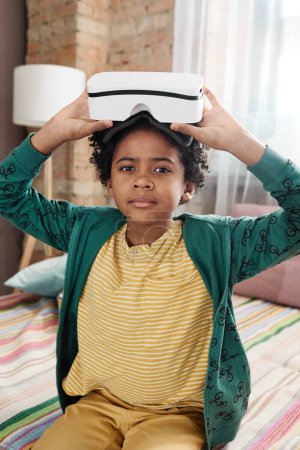 Photo for Portrait of African boy with curly hair looking at camera while wearing VR goggles on his head to play video game - Royalty Free Image