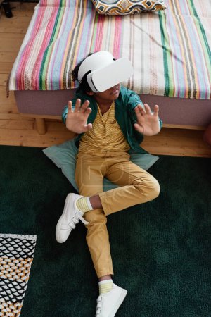 Photo for High angle view of African little boy in VR goggles sitting on floor and gesturing during virtual reality game - Royalty Free Image