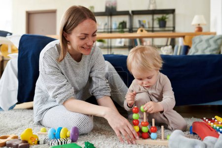 Sonriente joven madre sentada en el suelo y mirando por su pequeño hijo jugando juguetes educativos