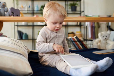 Photo for Cute blonde kid sitting on bed and touching the touch screen of digital tablet, he playing with it like a toy - Royalty Free Image