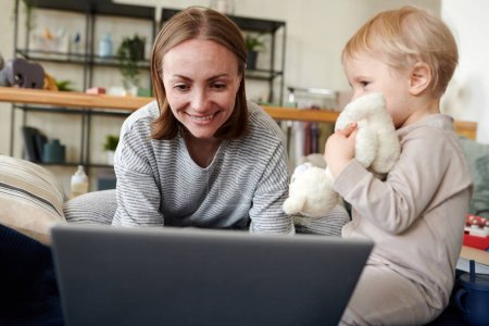Photo for Young smiling woman using her laptop while playing with her baby at home - Royalty Free Image