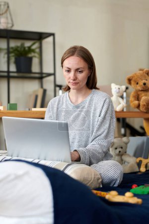 Photo for Young mother using laptop in her online work sitting on bed with toys during her maternity leave - Royalty Free Image