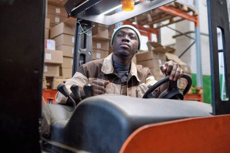 Serious African worker in uniform driving forklift at warehouse to load cargo