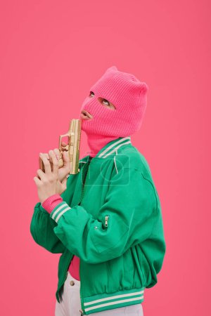 Junge Frau in pinkfarbener Sturmhaube und grüner Jacke posiert mit Pistole vor ihrem Gesicht auf rosa Hintergrund