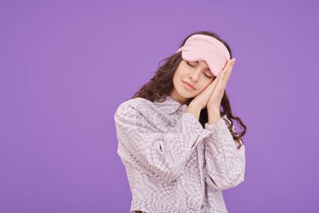 Portrait of young woman in sleep mask closed her eyes and putting head on her hands during dreaming against purple background
