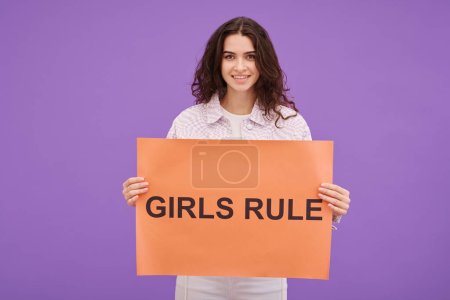 Photo for Portrait of young woman holding Girls Rule poster and smiling at camera against purple background - Royalty Free Image