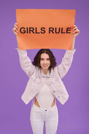 Photo for Portrait of young feminist girl holding poster above her head and speaking out standing against purple background - Royalty Free Image