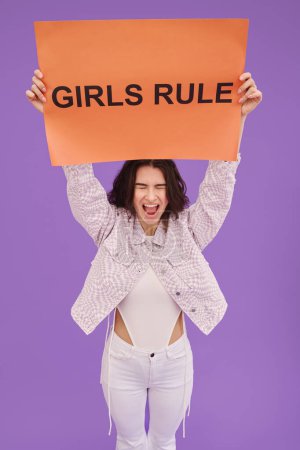 Photo for Portrait of young girl holding Girls rule poster above her head and screaming against purple background - Royalty Free Image