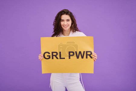 Photo for Portrait of young feminist girl with Girl Power placard smiling at camera standing on purple background - Royalty Free Image