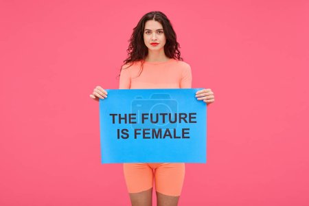 Photo for Portrait of young woman holding The future is female poster and looking at camera expressing her rights against pink background - Royalty Free Image