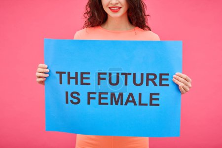 Photo for Close-up of young smiling woman holding The future is female poster to express her opinion in female rights isolated on pink background - Royalty Free Image