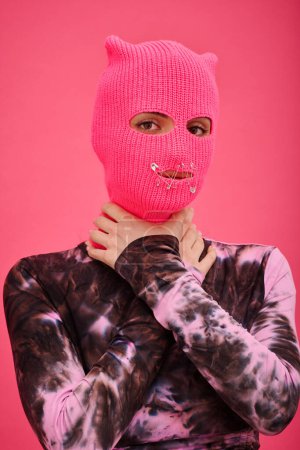 Photo for Portrait of young girl in balaclava with sewed mouth holding her neck and trying to speak out standing against pink background - Royalty Free Image