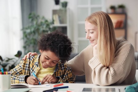 Foto de Joven madre sentada a la mesa en la habitación con su hijo adoptivo mientras él dibuja con lápices de colores - Imagen libre de derechos