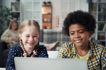 Téléchargez les photos : Enfants multiethniques regardant une vidéo drôle sur un ordinateur portable pendant que leur mère repose en arrière-plan - en image libre de droit