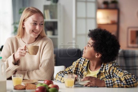 Foto de Joven madre feliz bebiendo café y hablando con su hijo adoptivo en la mesa durante el desayuno - Imagen libre de derechos