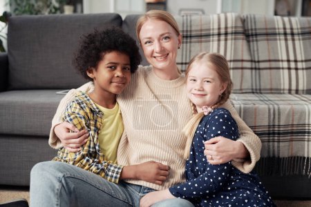 Retrato de la joven madre feliz abrazando a sus hijos adoptivos y sonriendo a la cámara sentada en el suelo en la habitación