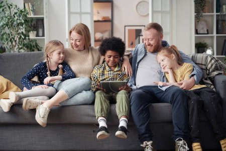 Niños adoptados viendo dibujos animados en tabletas digitales y disfrutando de pasar tiempo con padres adoptivos en el sofá en casa