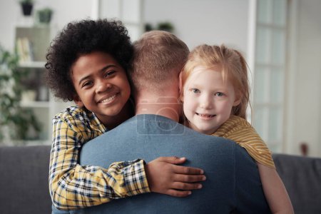Retrato de niños multiétnicos felices sonriendo a la cámara mientras abrazan a su padre adoptivo
