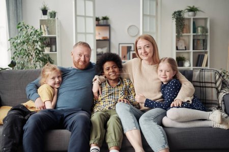 Portrait de famille adoptive heureuse avec leurs enfants embrassant ensemble et souriant à la caméra tout en se reposant sur le canapé dans le salon